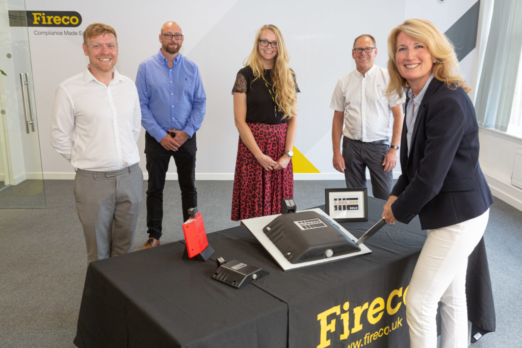Some of the Fireco team are standing around a cake. The cake was made to look like Dorgard, their door retaining device. They're all smiling stood around a table and the CEO is cutting the cake in celebration of selling one million Dorgards.