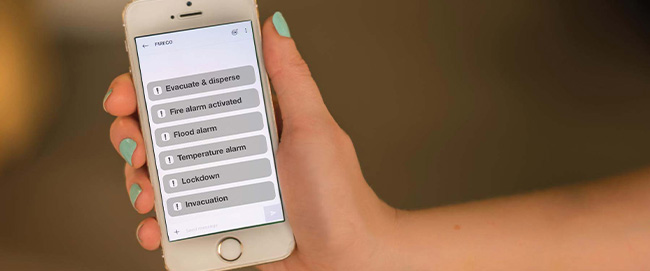 A hand with mint green nail varnish holding a white Iphone showing different alert types for Fireco's Digital Messaging Service (DMS).