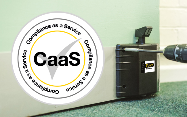 A black Dorgard SmartSound door retainer is being drilled onto the bottom of a green fire door. A CaaS Compliance as a Service logo sticker is on top of the photo.