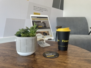 A round brown table hosts an ipad on a stand showing the fireco website, a plant, a coaster and a reusable coffee cup.
