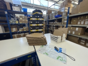 Two work benches are set up with lots of cardboard boxes and packing stacked up with shelves full of boxes in the background