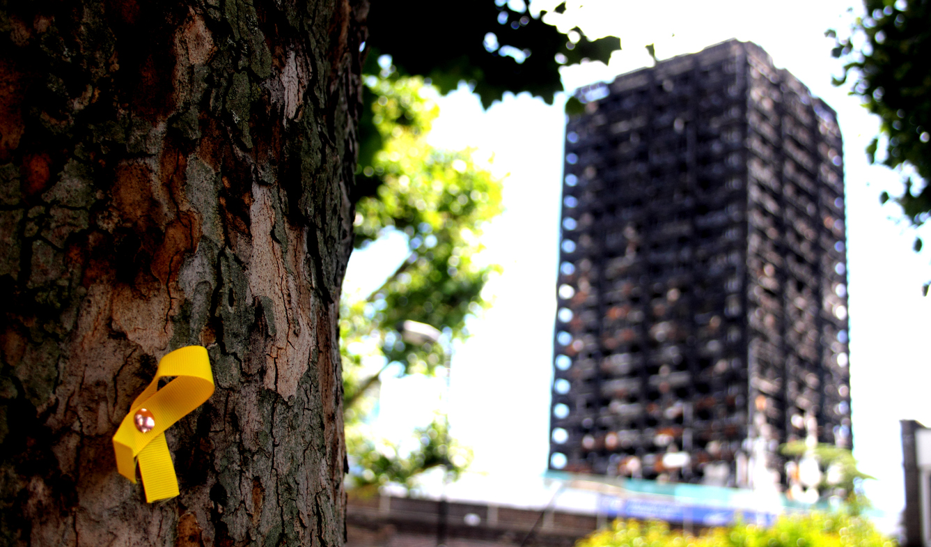 Burnt down Grenfell is visible in the background behind a tree with e yellow rememberance ribbon pinned on