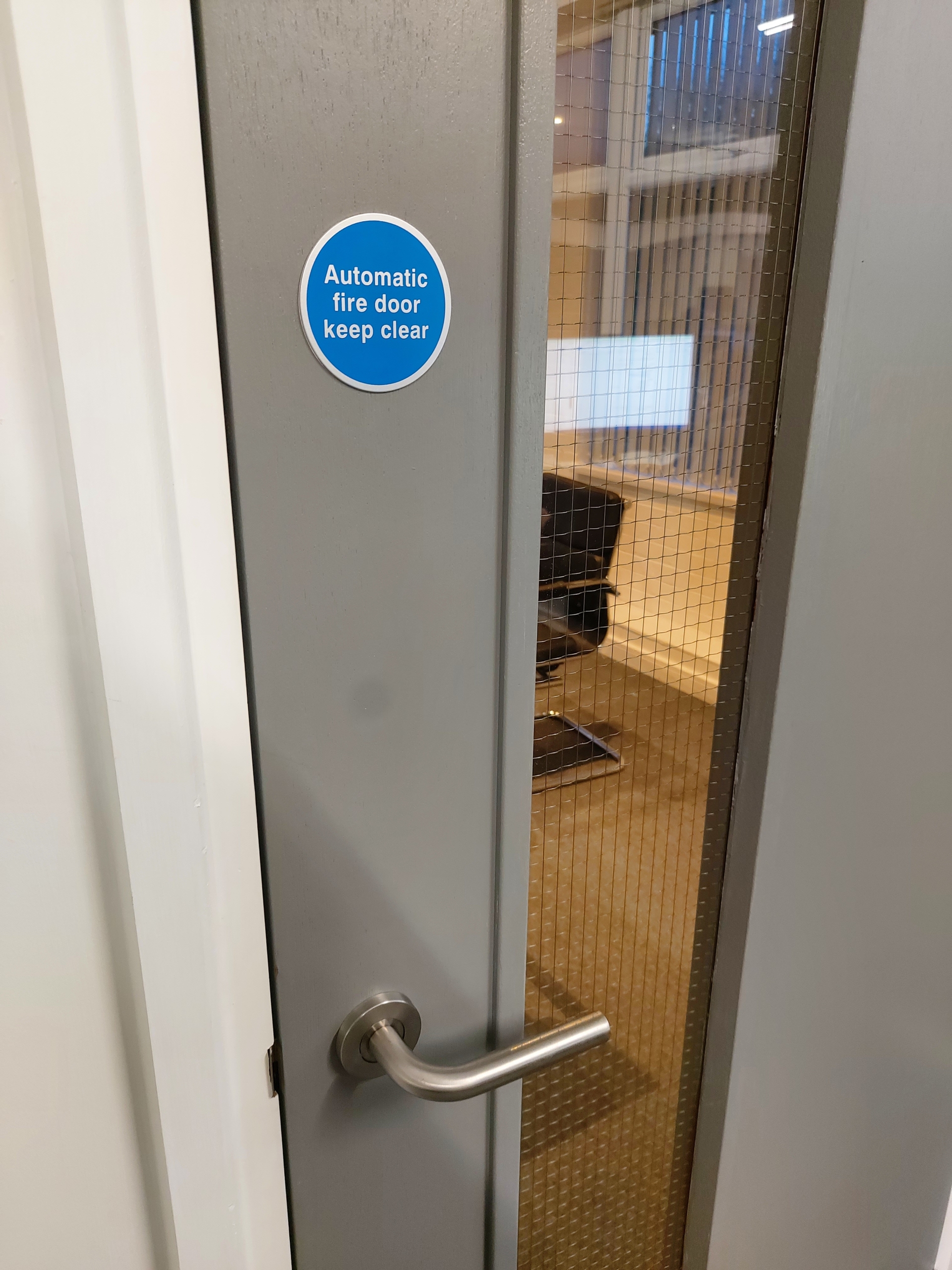 close up on a grey fire door with a blue circular sticker saying automatic fire door keep clear