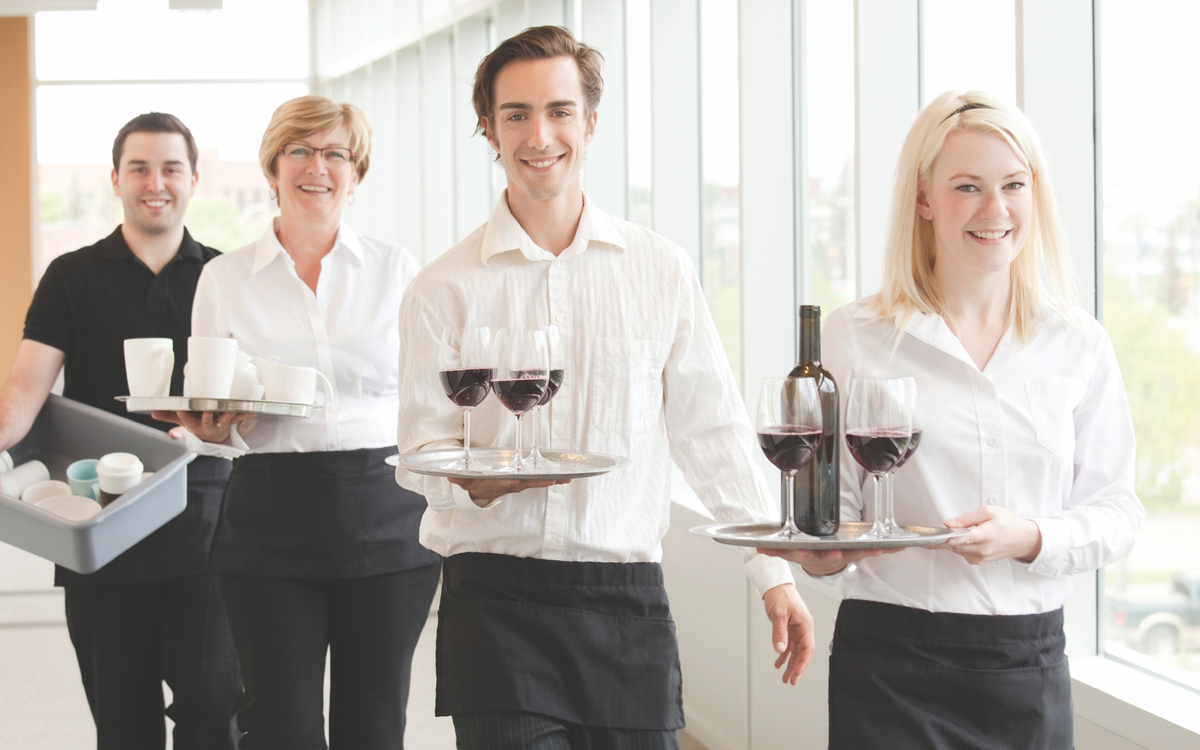 Hospitality staff walking in a line. They are each dressed smartly in white shirts and are carrying trays of drinks and wine bottles. Behind them follows a porter with a tray of dishes.