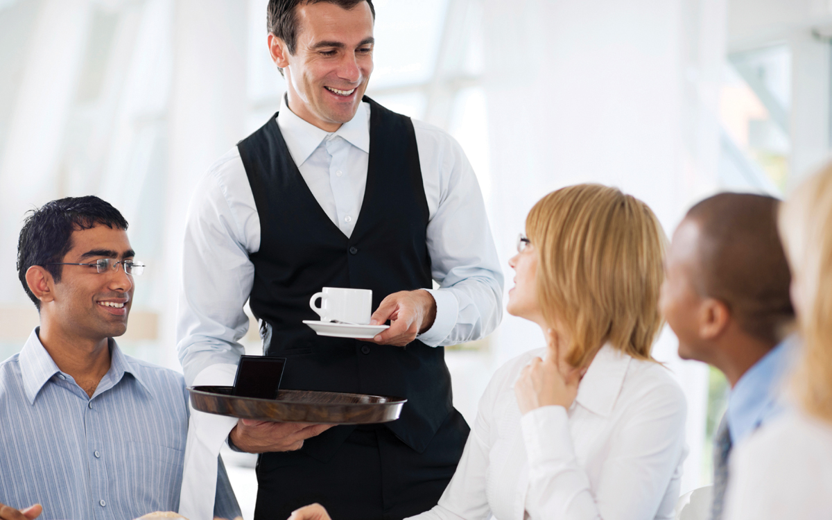 A waiter serving a table in a restaurant. The waiter is dressed smartly in a waistcoat and holds a tray of drinks.