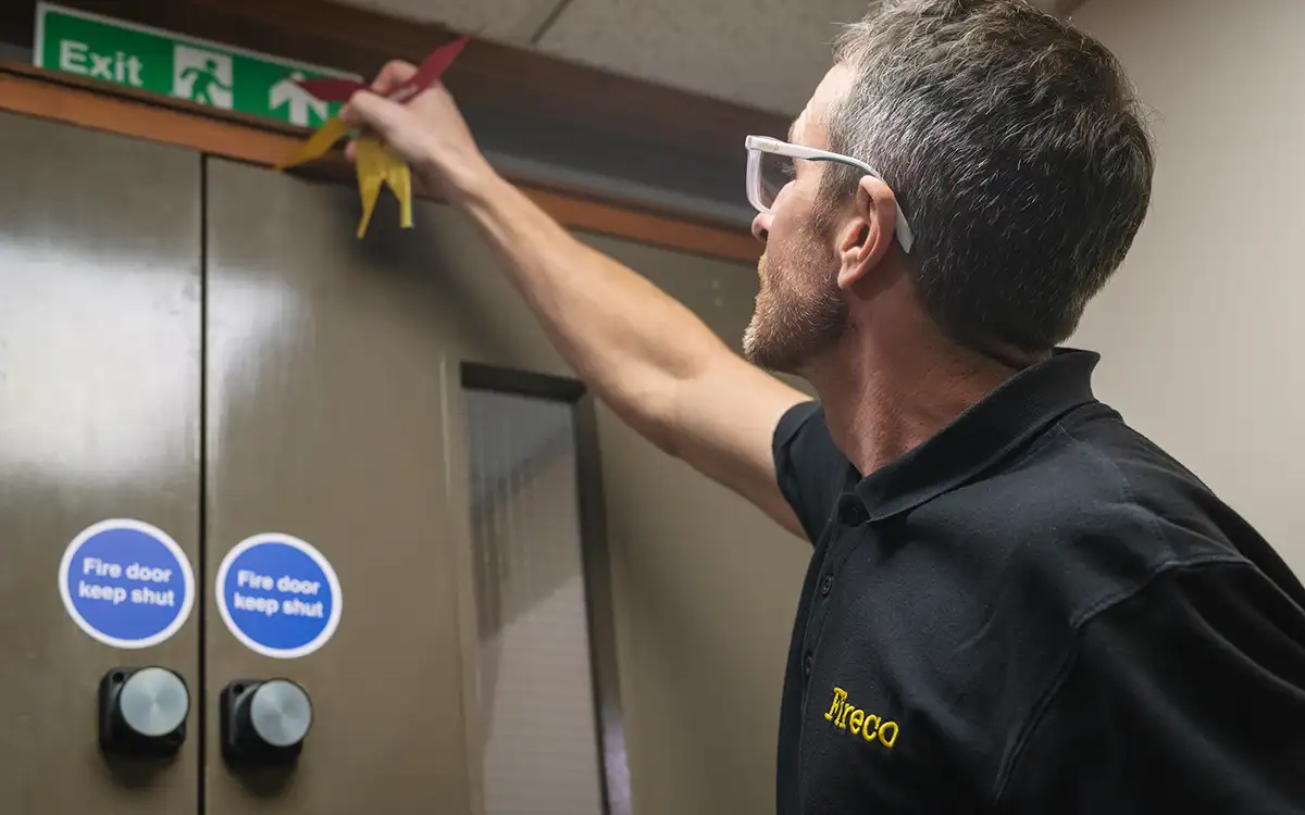An Engineer wearing a black t-shirt with the Fireco logo on is inspecting a fire door using a gap measurement tool
